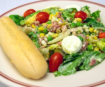 Close up of a cobb salad with romaine lettuce, grape tomatoes, feta cheese, corn, egg, and bacon, topped with a dressing and with a breadstick on the side
