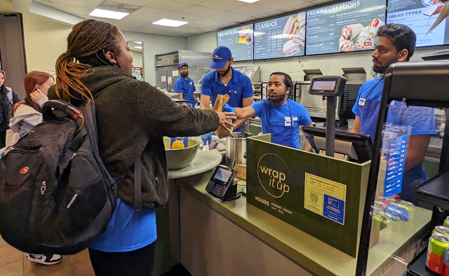 Associates handing a student their order; digital menuboards behind the associates