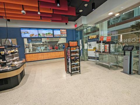 The interior of Corner Café at JSMBS. You can see some grab & go items, a snack shelf, the full digital menuboards above the associates preparing the food, a breakfast sandwich warmer, and one of the ordering kiosks.