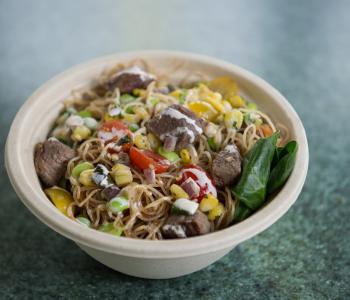 A bowl of noodles, corn, beef, edamame, and tomatoes, topped with a white sauce. Photo by Onion Studio.