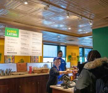 An associate cashing a student out after they order at the front of Sabor Latino. The printed menuboard is hanging from the ceiling in the background. Photo by Meredith Forrest Kulwicki.