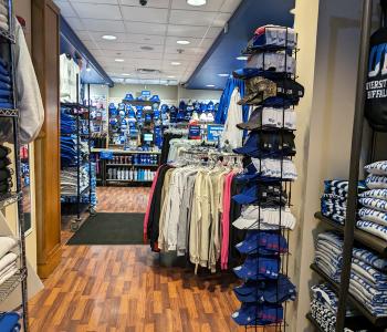 The small pathway within the Campus Tees store, facing the register where you can see hats and cups for sale. There are sweatshirts hanging on displays and also folded on shelves.