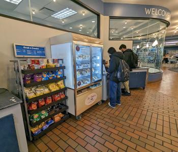 Snack shelves and bakery cabinet at Capen Café. Two students are preparing their bagels or getting a self serve cup of soup next to the cabinet.