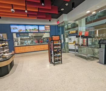 The interior of Corner Café at JSMBS. You can see some grab & go items, a snack shelf, the full digital menuboards above the associates preparing the food, a breakfast sandwich warmer, and one of the ordering kiosks.
