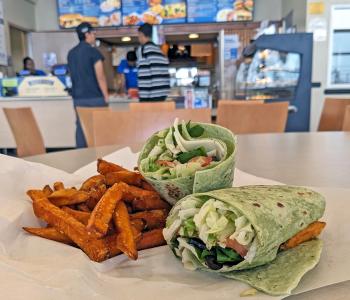 A veggie wrap and sweet potato fries on a table, with students ordering from Main Street Market in the background.