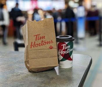A paper bag that says Tim Hortons, next to a Tim Hortons branded hot beverage cup, in front of the line at Tim Hortons in the Student Union.