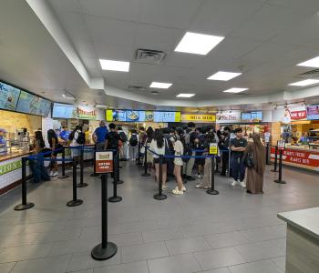 A view of Union Marketplace from the entrance, showing all stations from left to right: Union Station, Halal Shack, Piza Pizza, and Fowl Play. Customers are in line at every station.
