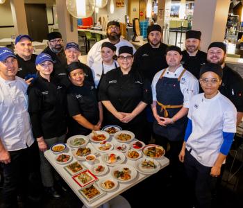 Executive Chef Amelia Ruiz (center) poses with the other chefs and staff responsible for the menu. Plated dishes from the event menu are placed in front of them on a table. Photo: Meredith Forrest Kulwicki